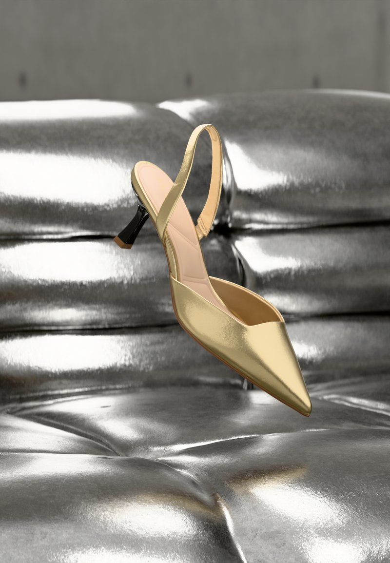 Gold pointed-toe slingback high heel shoe floating above a shiny metallic silver tufted leather couch.
