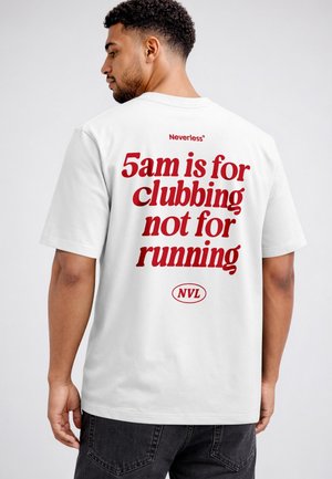Man with short curly hair wearing a white T-shirt with red text reading "5am is for clubbing not for running," standing and looking left.