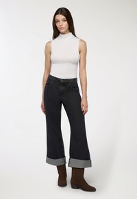 Donna in piedi con una canotta bianca senza maniche, jeans neri a gamba larga con risvolti e stivali alla caviglia marroni su uno sfondo bianco uniforme.