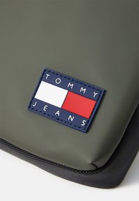 Grüne wasserdichte Tasche mit einem strukturierten schwarzen Reißverschluss, ausgestattet mit einem rechteckigen marineblauen Etikett mit dem Text "TOMMY JEANS" und rot-weiß-blauen Farbblöcken.