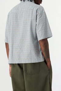 Homme portant une chemise à manches courtes à carreaux bleue et marron et un pantalon vert olive, les mains dans les poches, vu de dos.