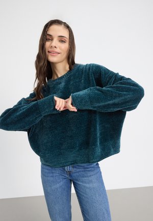 Tyrkis, plyssvettskjorte med brede ermer og ribbestrikkede cuffs. Kombinert med lyseblå jeans, mot en nøytral bakgrunn.