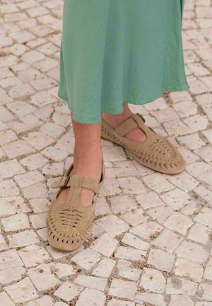 Des chaussures Mary Jane en suédine beige avec un design tissé et une bride à boucle, assorties à une robe verte fluide sur un sol en pavés.