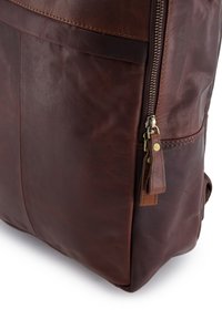 Brun læderrygsæk med en tekstureret overflade, lynlås og lædertrækflap. Har synlige sømme og sidepaneler.
