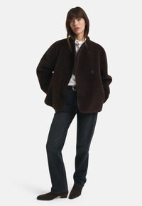 Žena v tmavo hnedej oversized sherpa bundě, bielej blúzke, čiernych džínsoch a tmavých členkových čižmách, stojí so svojimi rukami vo vreckách bundy.