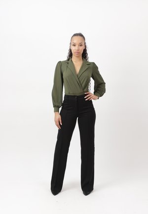 Vrouw staat zelfverzekerd, draagt een olijfgroene blouse met pofmouwen en zwarte hoge taille broek tegen een effen witte achtergrond.