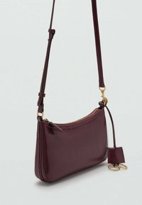 Geantă din piele bordeaux, tip crossbody, cu accesorii aurii, închidere cu fermoar, formă plată, curea ajustabilă și etichetă cu brelocuri atașate.