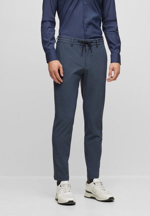 Dark blue tapered trousers made of textured fabric, featuring a drawstring waist and two side pockets, paired with white sneakers.