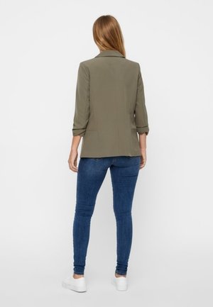 Femme debout, vue de dos, portant un blazer vert olive, un jean skinny bleu et des baskets blanches sur un fond blanc uni.