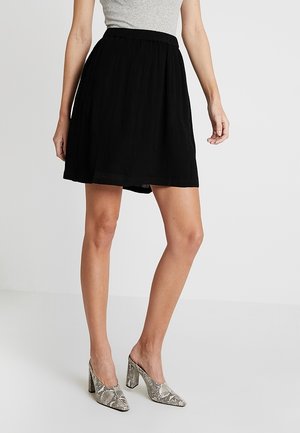 Frau trägt einen schwarzen kniehohen Rock, graues ärmelloses Oberteil und hochhackige Schuhe mit Schlangenledermuster, steht vor einem einfarbigen Hintergrund.