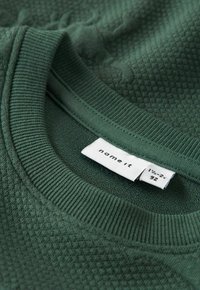 Sweatshirt vert en tissu texturé avec un col côtelé. L'étiquette indique la taille 1½-2 ans. Le tissu présente un subtil motif en losange.