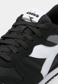 Black sneaker with white accents, textured fabric upper, and round toe. Features flat laces and a logo on the tongue. Rubber sole.