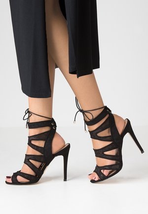 Voeten in zwarte strappy high heels met enkelbandjes, gecombineerd met zwarte cropped broeken tegen een neutrale lichte achtergrond.
