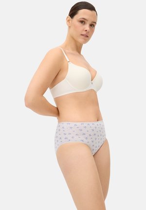 Mujer con un sujetador blanco y bragas altas de color púrpura claro con un pequeño estampado floral, de pie con las manos detrás de la espalda sobre un fondo liso.