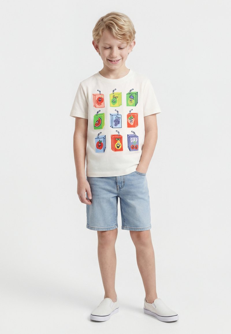 Ξανθός αγόρι που χαμογελά, φοράει λευκό μπλουζάκι με πολύχρωμα σκίτσα κουτιών χυμού, ανοιχτόχρωμο τζιν σορτς και λευκά παπούτσια τύπου slip-on, στέκεται σε λευκό φόντο.