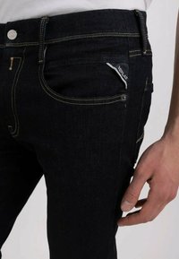 Jeans en denim bleu foncé avec des coutures jaunes, une petite poche à monnaie et un matériel métallique au niveau de la taille. Texture lisse avec un léger éclat.