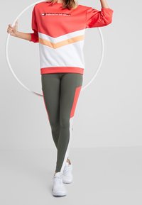 Sudadera roja y blanca con patrón de chevron naranja; leggings verdes con acentos rojos y blancos; modelo sostiene un aro; zapatillas deportivas.
