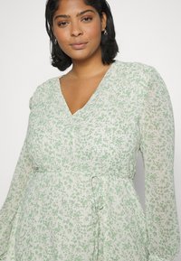 Vestido leve de mangas longas em branco com estampado floral verde, apresentando um decote em V e cintura com laço. Tecido fluido com uma textura suave.