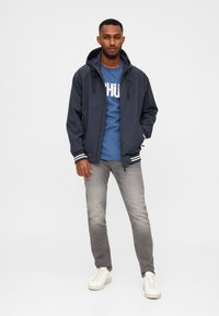 Giacca a vento navy con cappuccio, strisce bianche sui polsini, abbinata a una maglietta blu con testo bianco in grassetto e jeans grigi. Sneakers bianche.