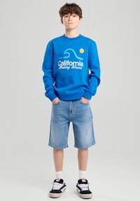 Sininen huppari, jossa on teksti "California Making Waves" ja aaltojen grafiikka. Yhdistetty vaaleisiin farkkushortseihin ja mustiin lenkkitossuihin, joissa on valkoisia yksityiskohtia.