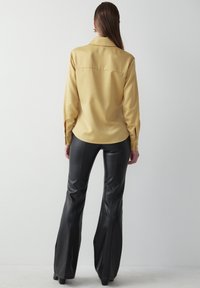 Langærmet gul skjorte lavet af glat stof, parret med sorte, flagrende vinylbukser. Skjorten har en krave og knapmanchetter.