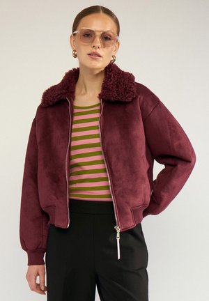 Frau trägt transparente rosa Sonnenbrille, goldene Creolen, weinrote Jacke mit Lammfellkragen, gestreiftes grün-rosa Hemd und schwarze Hose.