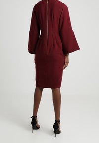 Viininpunainen vartalonmyötäinen mekko, jossa liehuvat pitkät hihat, takana vetoketjusomiste ja polvipituinen leikkaus; sileä, jämäkkä kangaspinta.