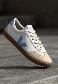 Sneaker bianchi e beige con accenti blu chiaro, caratterizzati da un design con lacci, punta in suede e suola in gomma con superficie texturizzata.