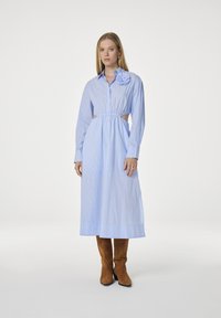 Lichtblauwe gestreepte shirtjurk met lange mouwen, uitsnijdingen bij de taille en een bloemaccent op de schouder. Draag met een paar bruine suède laarzen.