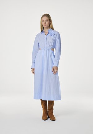 Lichtblauwe gestreepte shirtjurk met lange mouwen, uitsnijdingen bij de taille en een bloemaccent op de schouder. Draag met een paar bruine suède laarzen.