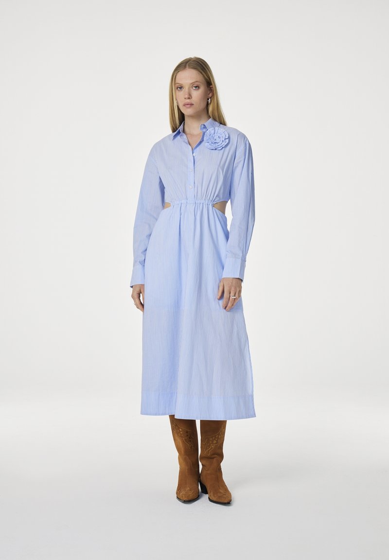 Lichtblauwe gestreepte shirtjurk met lange mouwen, uitsnijdingen bij de taille en een bloemaccent op de schouder. Draag met een paar bruine suède laarzen.