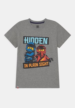 Graues T-Shirt mit zwei LEGO-Ninja-Figuren, eine in Blau und eine in Rot, mit dem Text "HIDDEN IN PLAIN SIGHT."