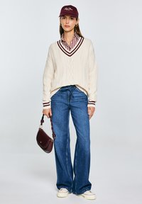 Cremfarbener Zopfstrickpullover mit maroonfarbenen Akzenten, gestreiftem Kragenhemd, weiten blauen Jeans und einer maroonfarbenen Mütze. Hält eine weinrote Handtasche.