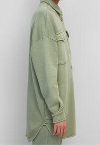 Persoon in een oversized lichtgroene button-up jas met borstzakken, zijsplithem en bijpassende broek tegen een effen achtergrond.