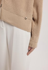 Beżowy sweter z dzianiny o wzorzystej fakturze, z prążkowymi mankietami i srebrnym guzikiem, zestawiony z białymi spodniami o szerokich nogawkach.