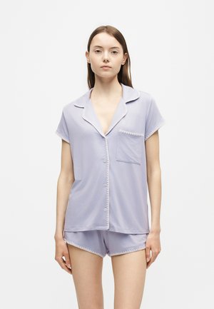 Joven mujer que lleva una camiseta de pijama de manga corta y color lila claro con botones y pantalones cortos a juego, de pie frente a un fondo blanco liso.