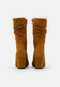 Bottines en daim marron haute jusqu'au genou avec un design décontracté, dotées d'un talon block et d'une fermeture éclair sur le côté pour un enfilage facile. Texture lisse, détails minimaux.
