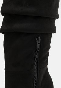 Svart semsket støvel med en slouchy overdel, som har glidelås på siden. Den glatte teksturen og den tettsittende formen fremhever designet.