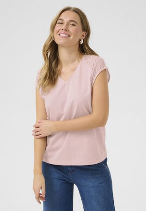 T-shirt rose pâle avec un col en V, des manches courtes en dentelle et une coupe décontractée. Porté avec un jean bleu, au toucher lisse.