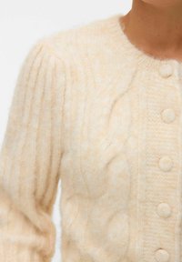 Ljus beige stickad cardigan med kabelmönster, rund hals och sex texturerade knappar fram. Mjuk, fluffig textur.