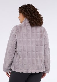 Kurtka z futerka ekologicznego w jasnoszarym kolorze o pikowanej fakturze, z wysokim kołnierzem i falowanym dołem. Posiada długie rękawy i jest noszona na ciemnych spodniach.
