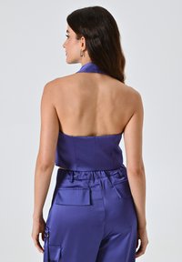 Top corto a halter in satin viola con un design aderente, che mette in risalto la schiena. Abbinato a pantaloni in satin coordinati con tasche.
