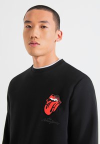 Sudadera negra con el logotipo bordado de los Rolling Stones, con labios rojos y lengua. Cuello redondo y tejido texturizado, diseño ajustado.