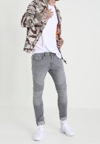 Chaqueta bomber de camuflaje con jeans grises que presentan detalles acanalados en las rodillas y una camiseta blanca; combinada con zapatillas blancas.