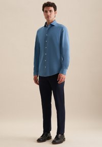 Uomo in piedi che indossa una camicia azzurra con bottoni a maniche lunghe, pantaloni scuri navy e scarpe eleganti nere, su uno sfondo neutro.