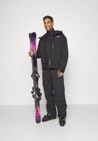 Black ski jacket and pants, with a glossy finish. Holds purple and black skis featuring a gradient design and bindings.