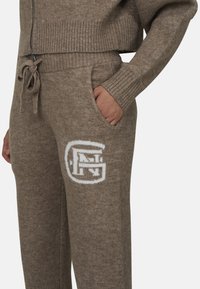 Brązowe dzianinowe spodnie dresowe ze sznurkiem w talii, bocznymi kieszeniami oraz białym logo na lewej udzie. Teksturowany, miękki materiał.