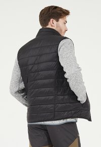 Vadderad svart väst med hög krage, med horisontella quiltade sektioner. Bärs över en ljusgrå långärmad topp.