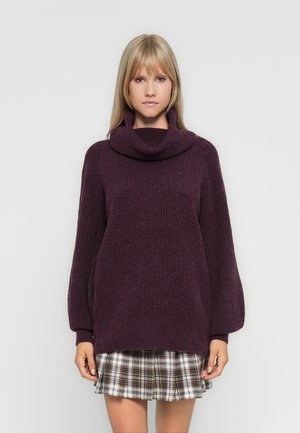 Pull en violet tricoté avec un col roulé haut, des manches surdimensionnées et une texture côtelée, associé à une mini jupe à carreaux.