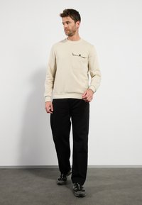Lichtbeige sweatshirt met een ronde hals, voorzien van een borstzakje aan de linkerkant en een knop. Draagt bij zwarte broek en sportschoenen.